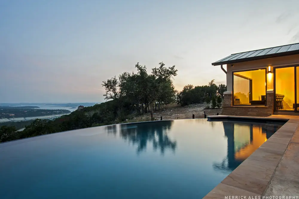 Front elevation at dusk of a contemporary stone and stucco home with a metal roof and lit garage bays across a wide drive.