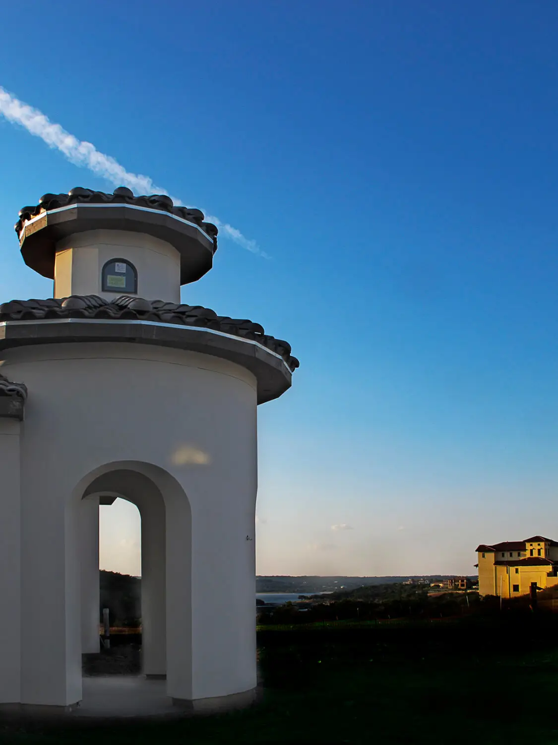 Front elevation at dusk of a Mediterranean home with a stucco turret, tile roof and lit arched entry above a curved drive.