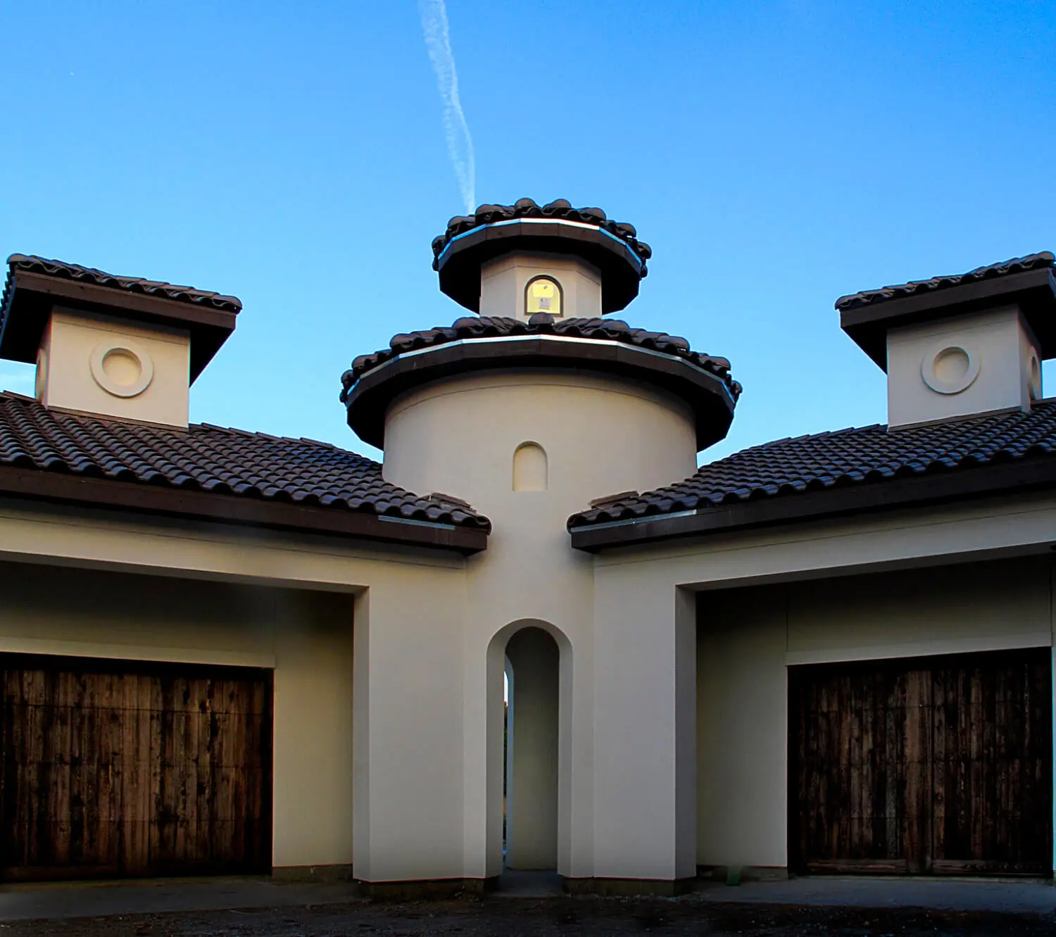 Front elevation at dusk of a Mediterranean home with a stucco turret, tile roof and lit arched entry above a curved drive.
