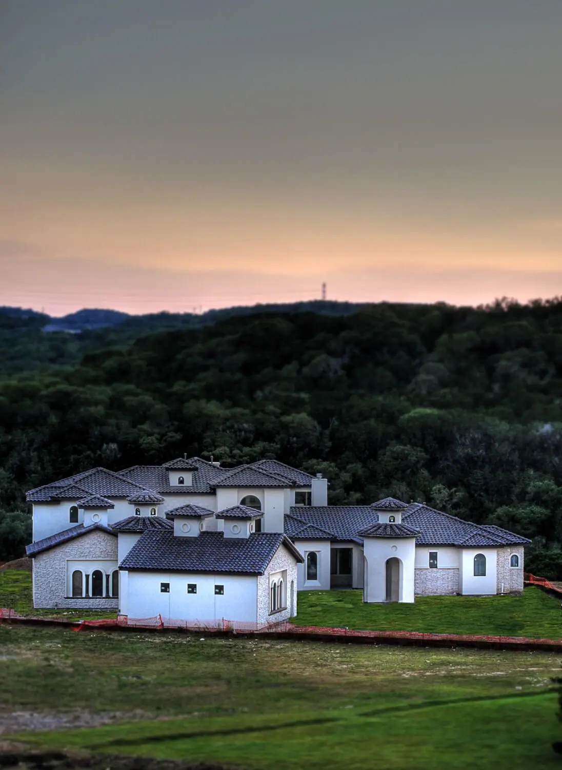 Front elevation at dusk of a Mediterranean home with a stucco turret, tile roof and lit arched entry above a curved drive.
