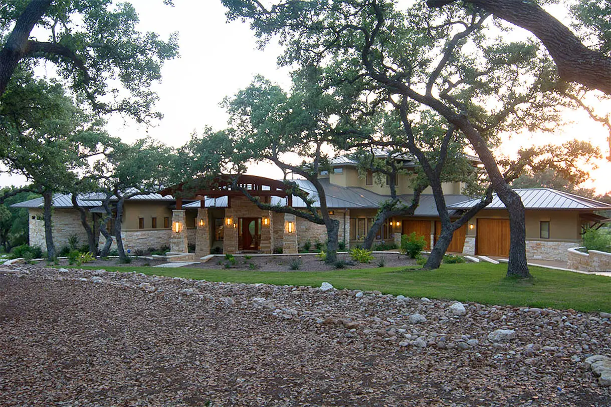 Long stone home with a curved timber entry canopy, seen at dusk through the trunks of live oaks over a gravel bed.