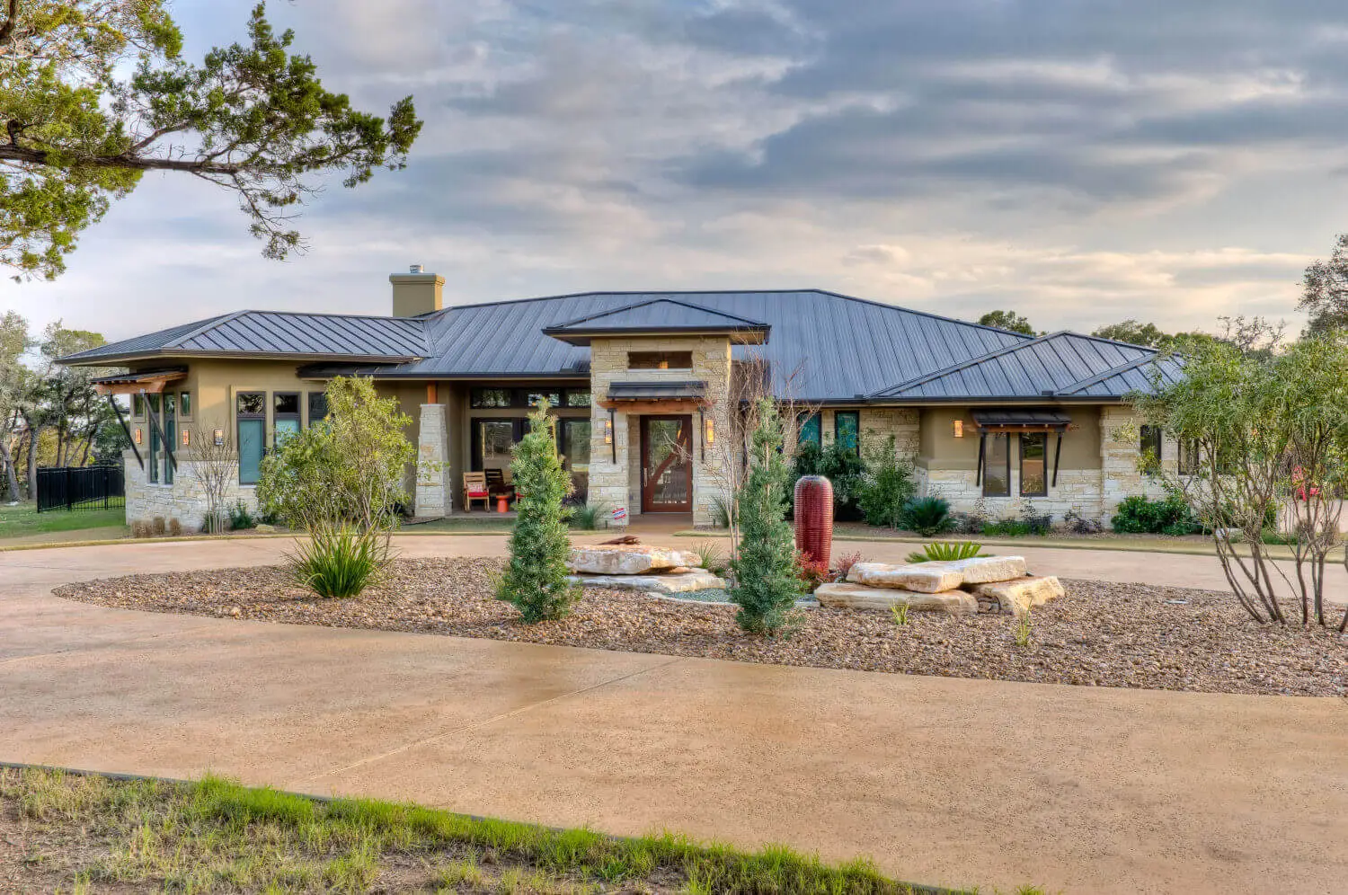 Low stone home with a standing seam metal roof and a recessed entry, seen across a gravel motor court under a clouded sky.