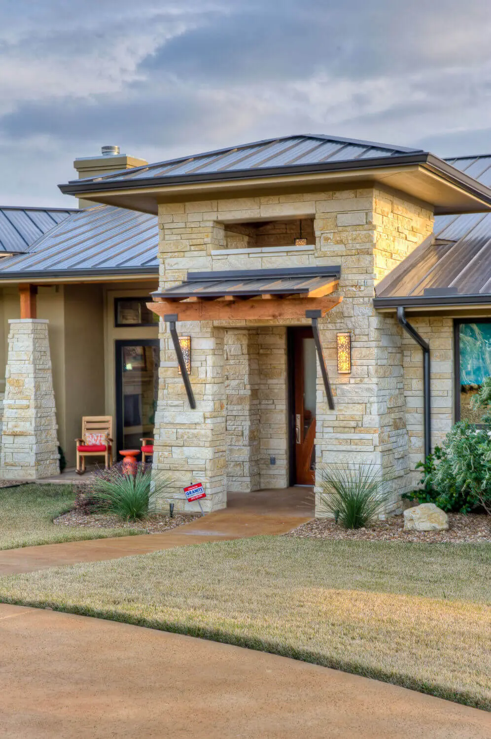 Long low home with a metal roof and stone base, seen across open ground between two oaks.