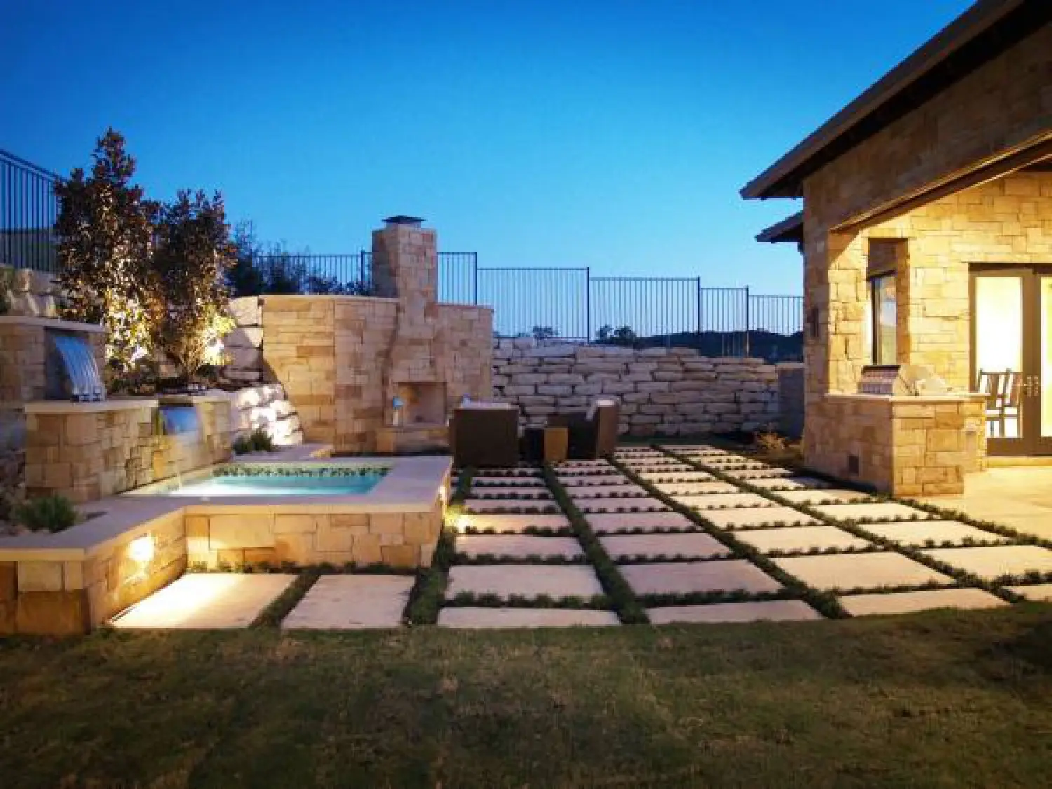 Stone entry elevation lit at dusk, with a shed-roofed porch, tall windows and a low wall along the walk.