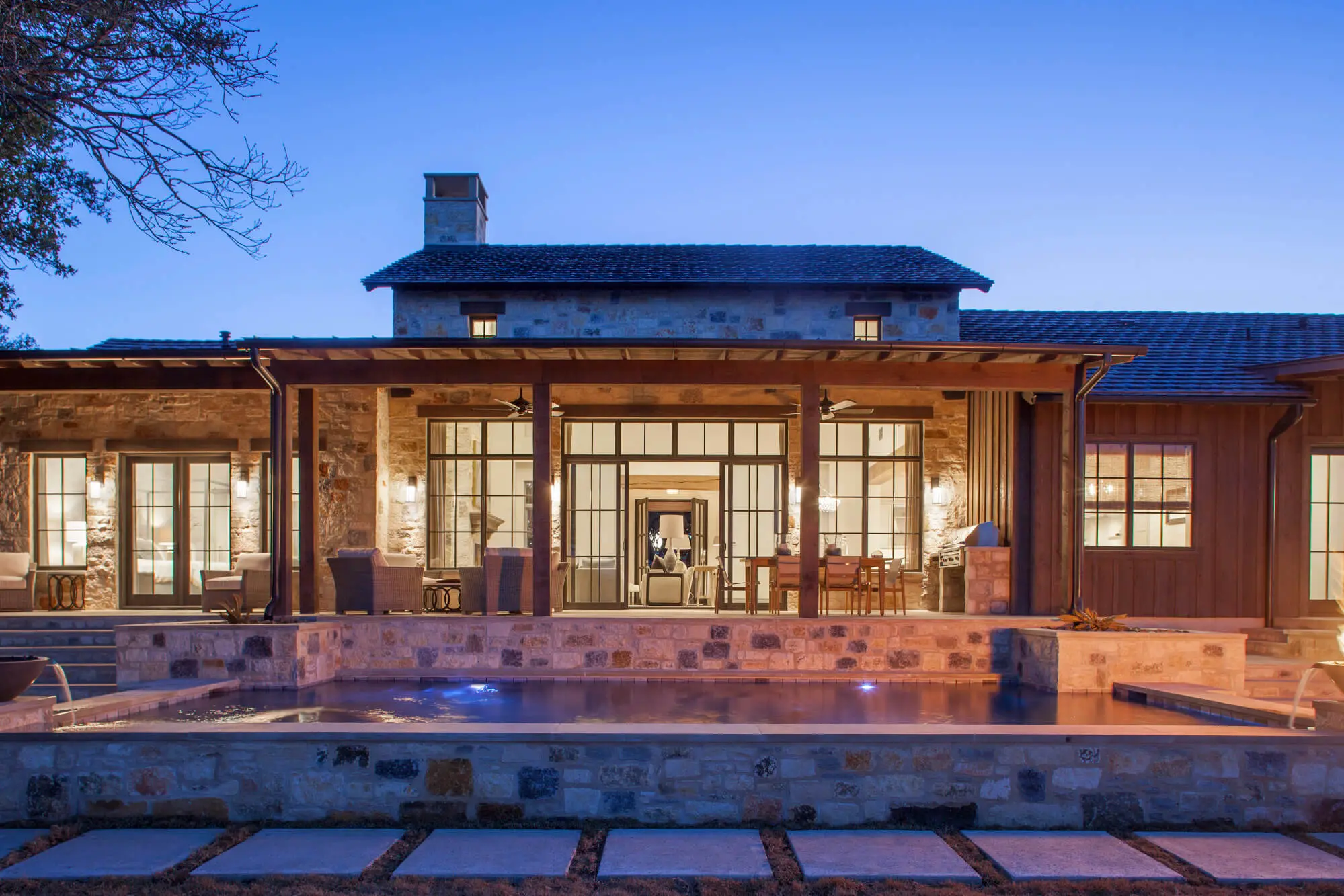 Rear elevation lit from within at dusk, with a deep covered porch running the length of the house above a stone-edged pool.