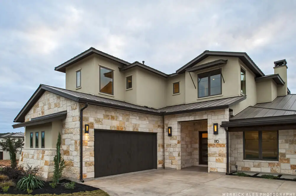 Front elevation of a two-story stone and stucco home with two garage bays and planted beds along the drive.
