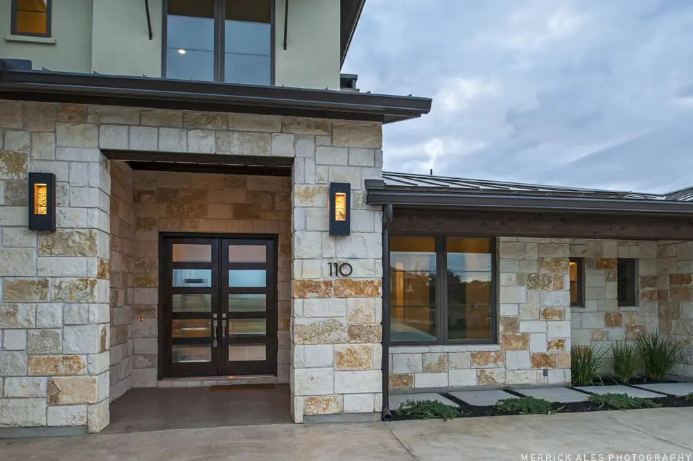 Front elevation of a two-story stone and stucco home with two garage bays and planted beds along the drive.
