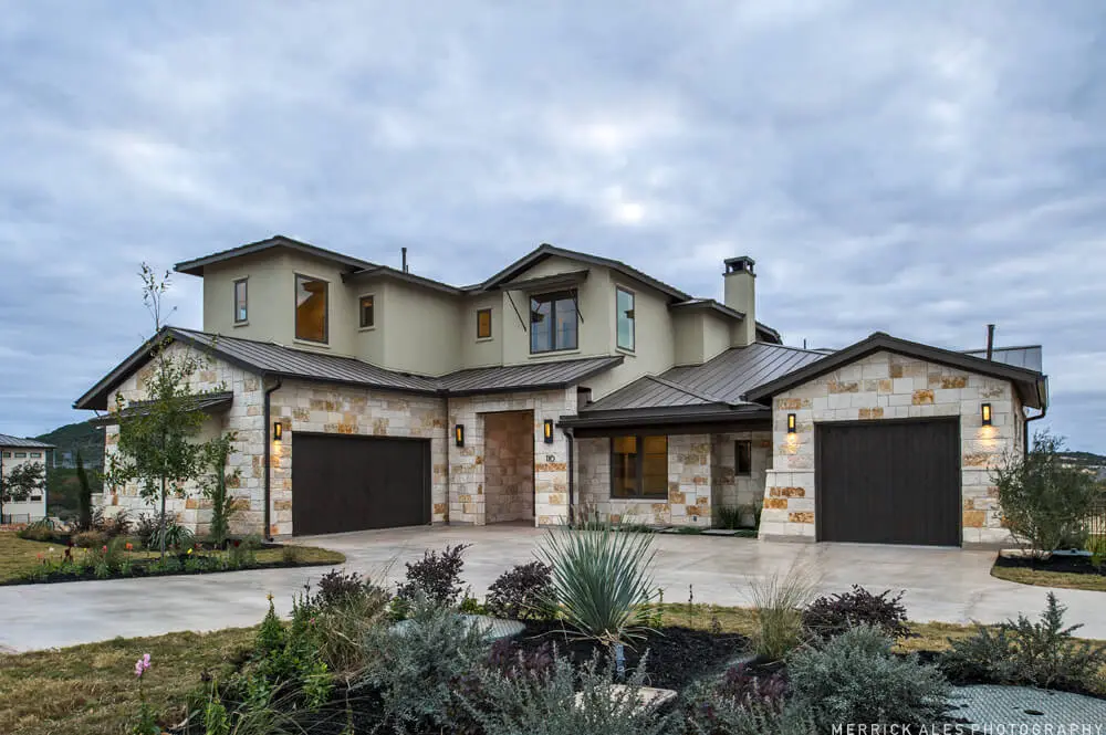 Front elevation of a two-story stone and stucco home with two garage bays and planted beds along the drive.