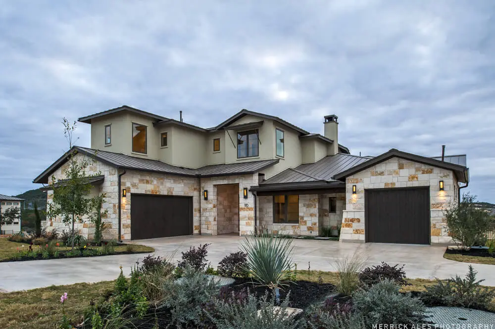 Front elevation of a two-storey stone and stucco house with two garage bays and planted beds along the drive.