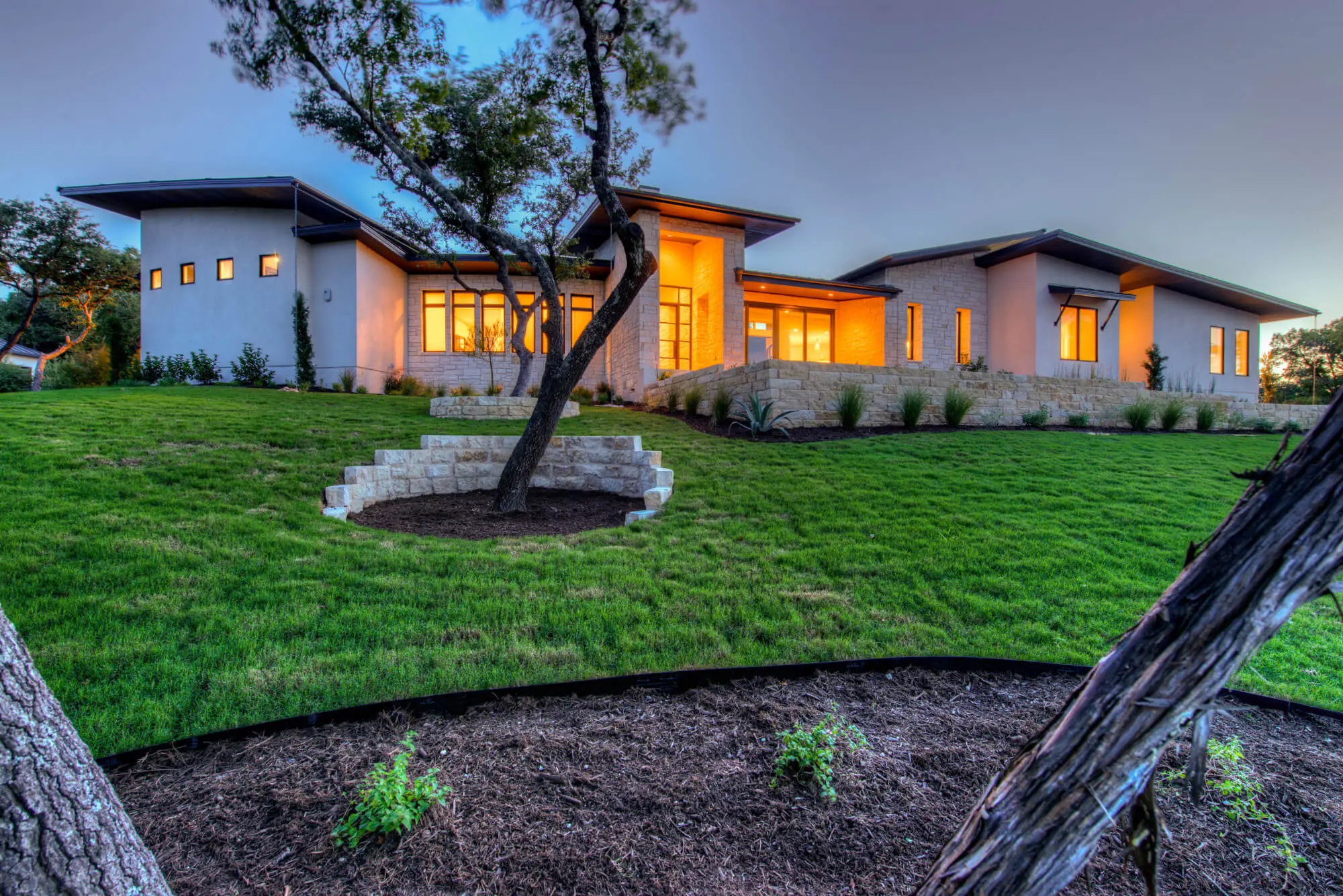 A low modern home lit from within at dusk, its wide overhanging roofs stepping down a planted slope.
