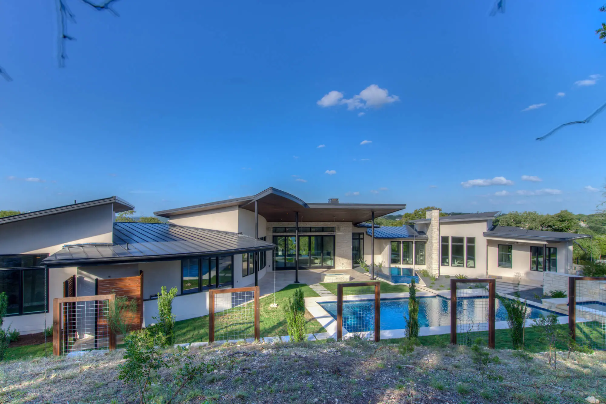 Rear elevation of a low modern house with a wide overhanging roof, glass walls and a pool terrace, seen from the slope below.
