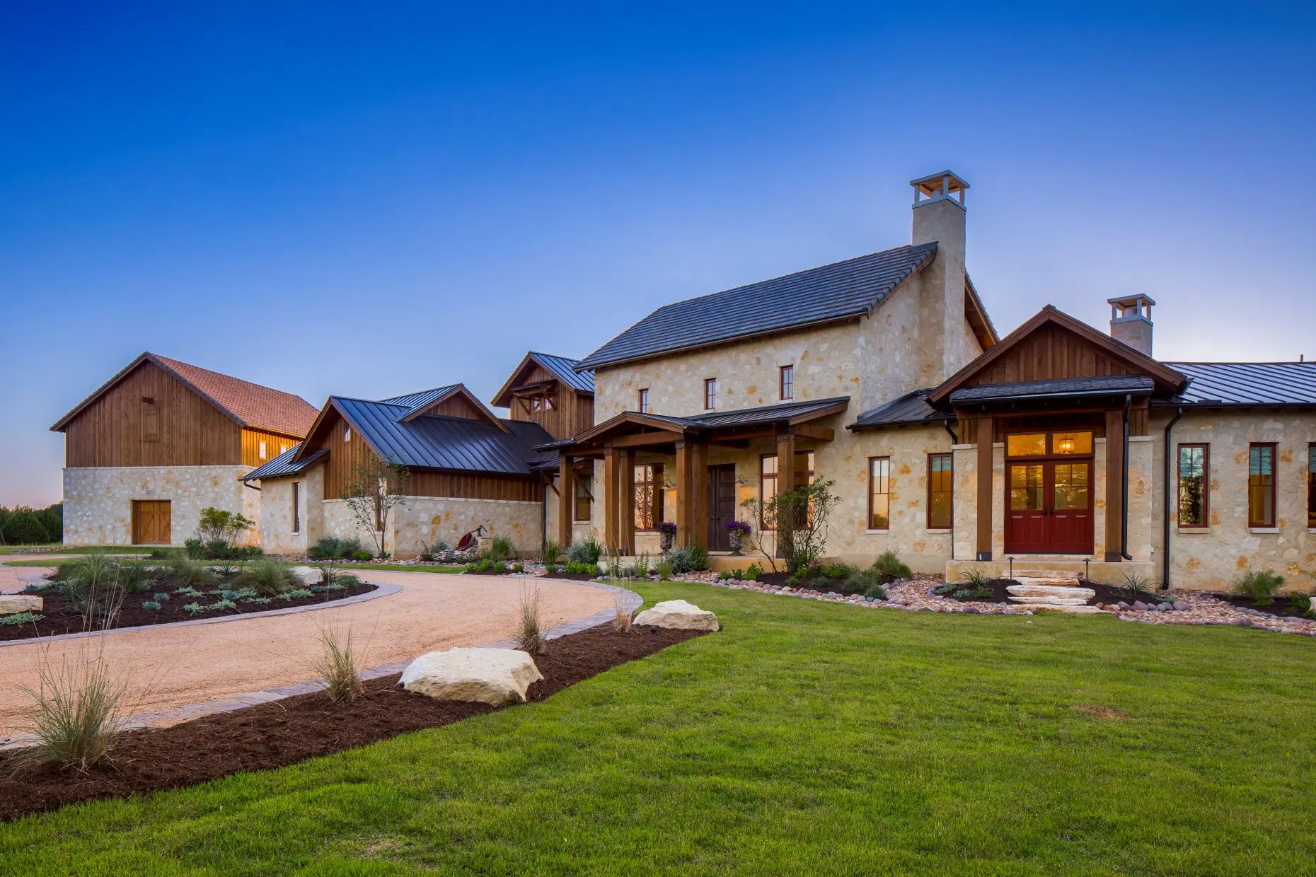 Front elevation at dusk of a stone farmhouse with metal roofs and a red front door, across a broad lawn and gravel drive.