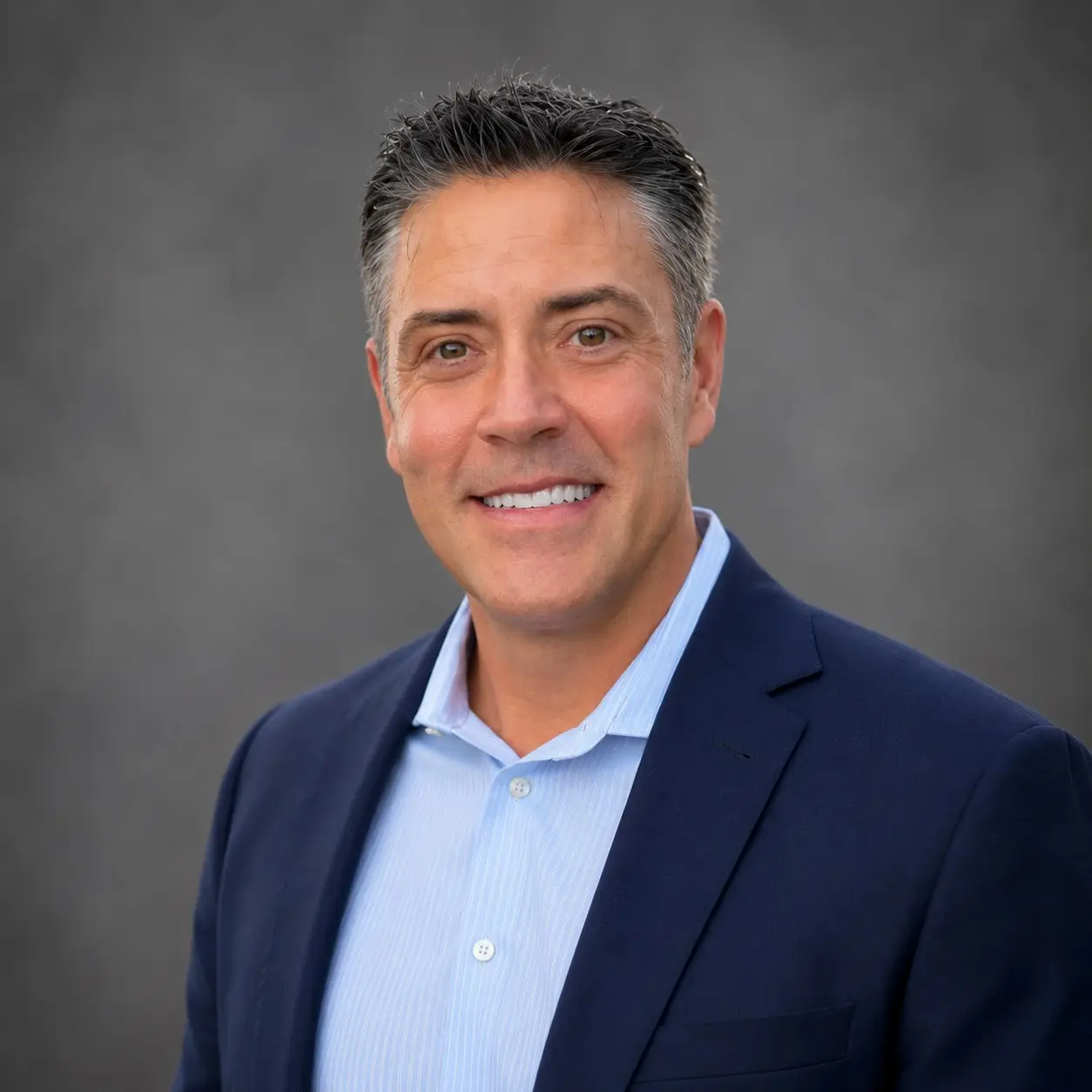 Curtis Raymond, a founding partner of the studio, in a navy jacket and open-necked striped shirt against a grey studio background.