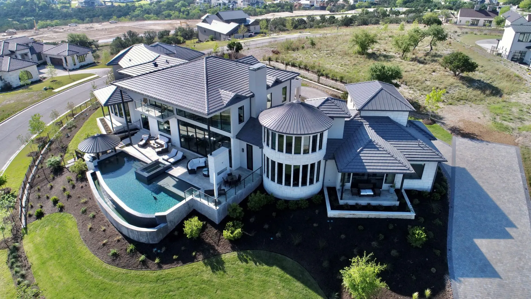 Aerial view of a house with a round tower and a curved pool terrace, on a lawn edged with new planting.