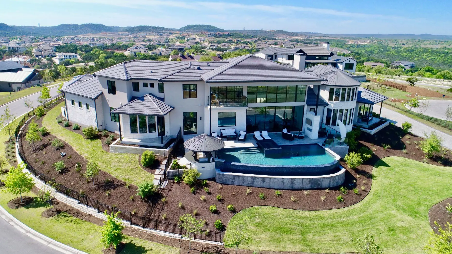 Aerial view of a home with a round tower and a curved pool terrace, on a lawn edged with new planting.