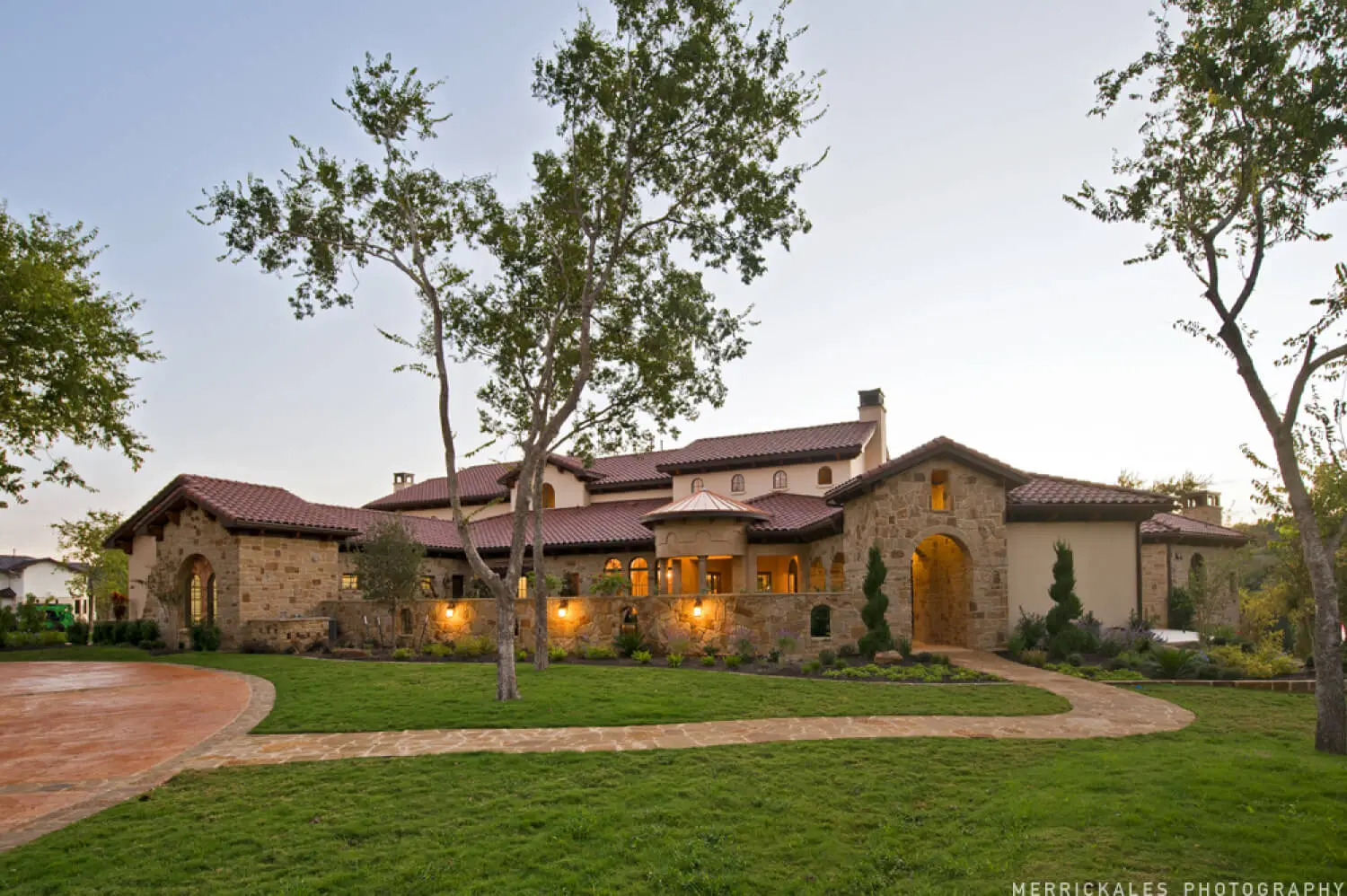 Front elevation at dusk of a stone and stucco home with a tile roof, lit entry arch and a flagstone walk across the lawn.
