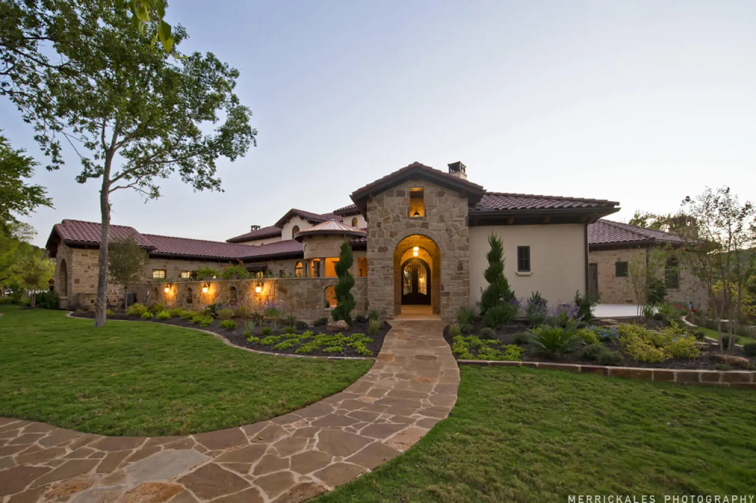 Front elevation at dusk of a stone and stucco house with a tile roof, lit entry arch and a flagstone walk across the lawn.