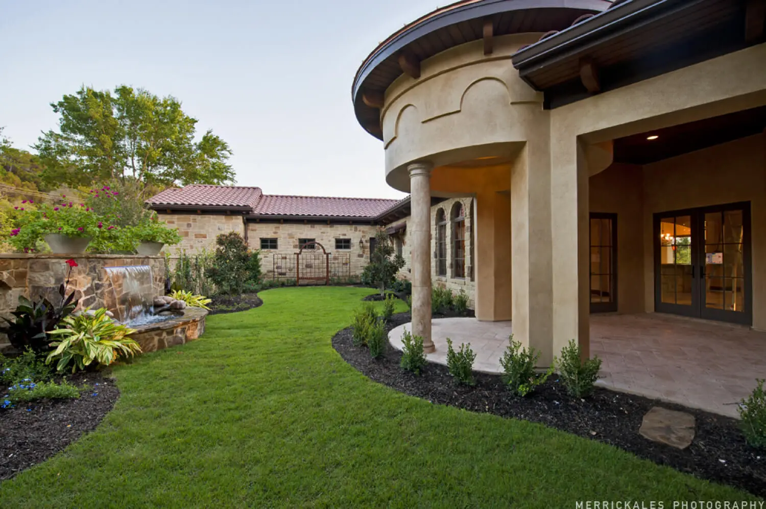 Front elevation at dusk of a stone and stucco home with a tile roof, lit entry arch and a flagstone walk across the lawn.