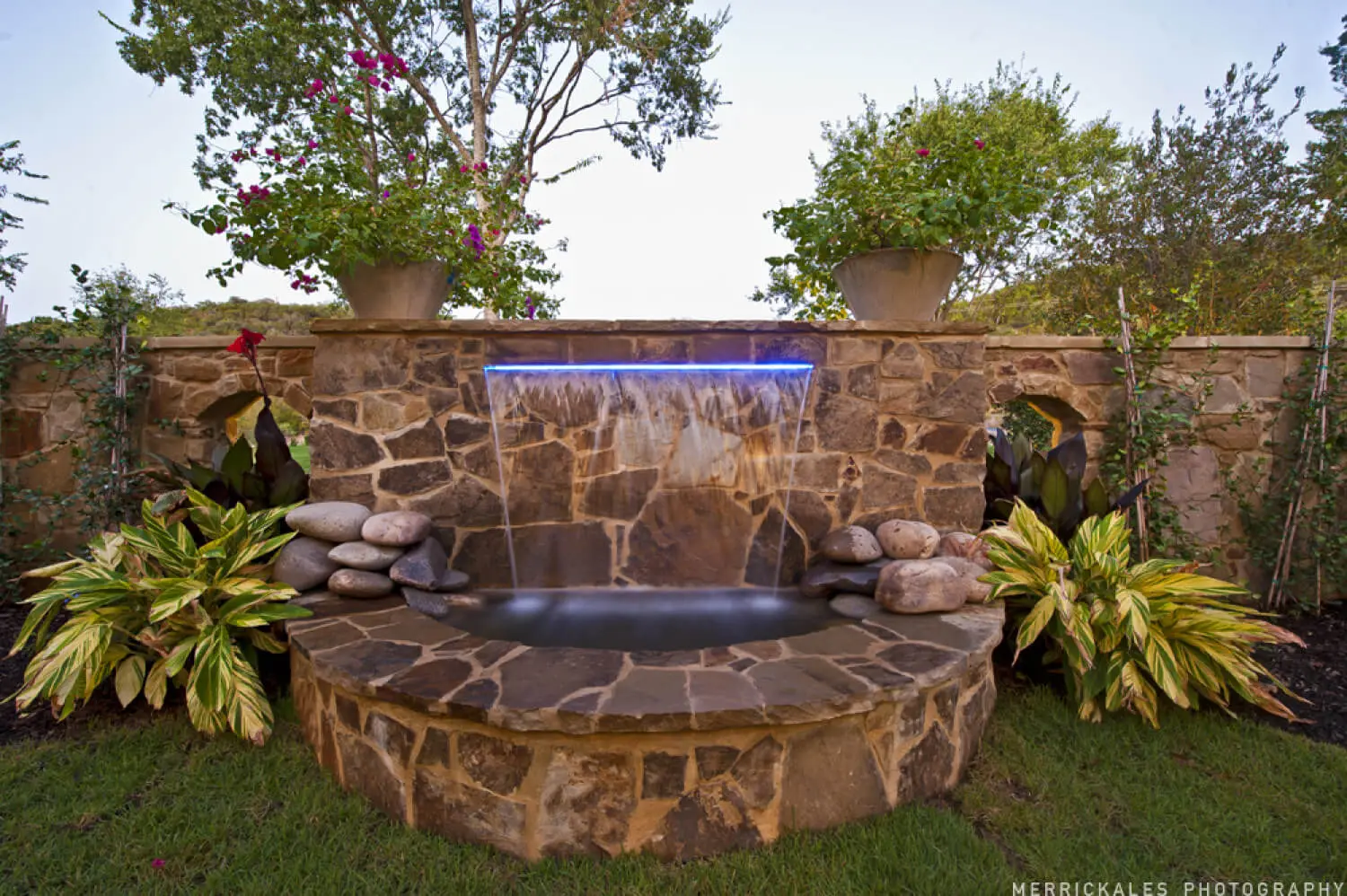 Front elevation at dusk of a stone and stucco home with a tile roof, lit entry arch and a flagstone walk across the lawn.