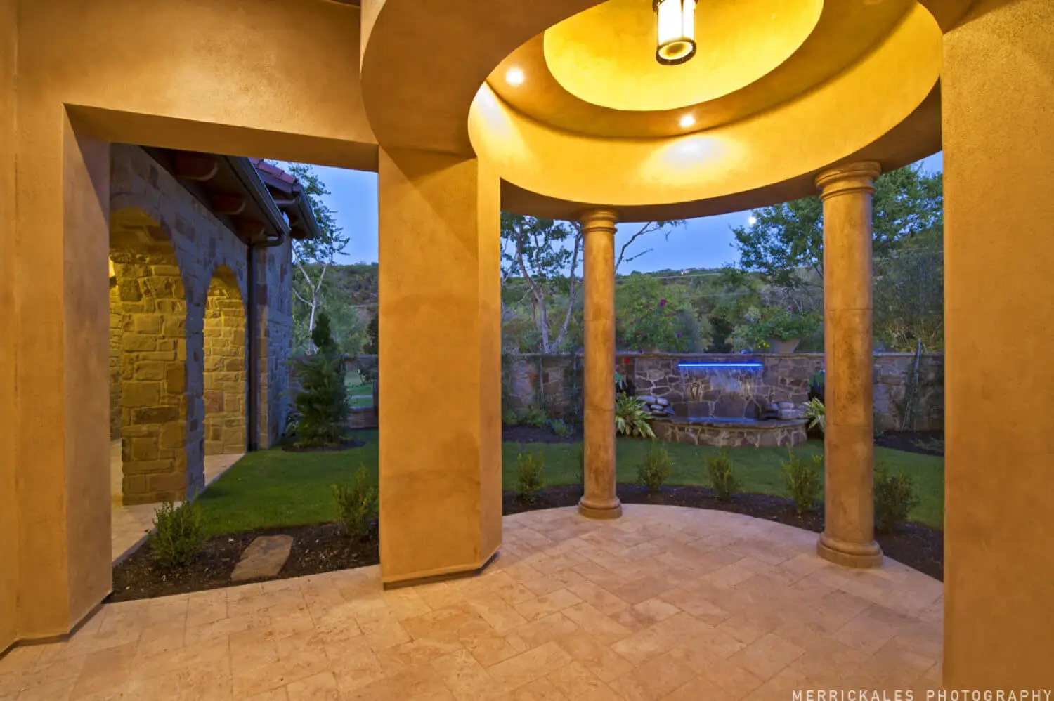 Front elevation at dusk of a stone and stucco home with a tile roof, lit entry arch and a flagstone walk across the lawn.