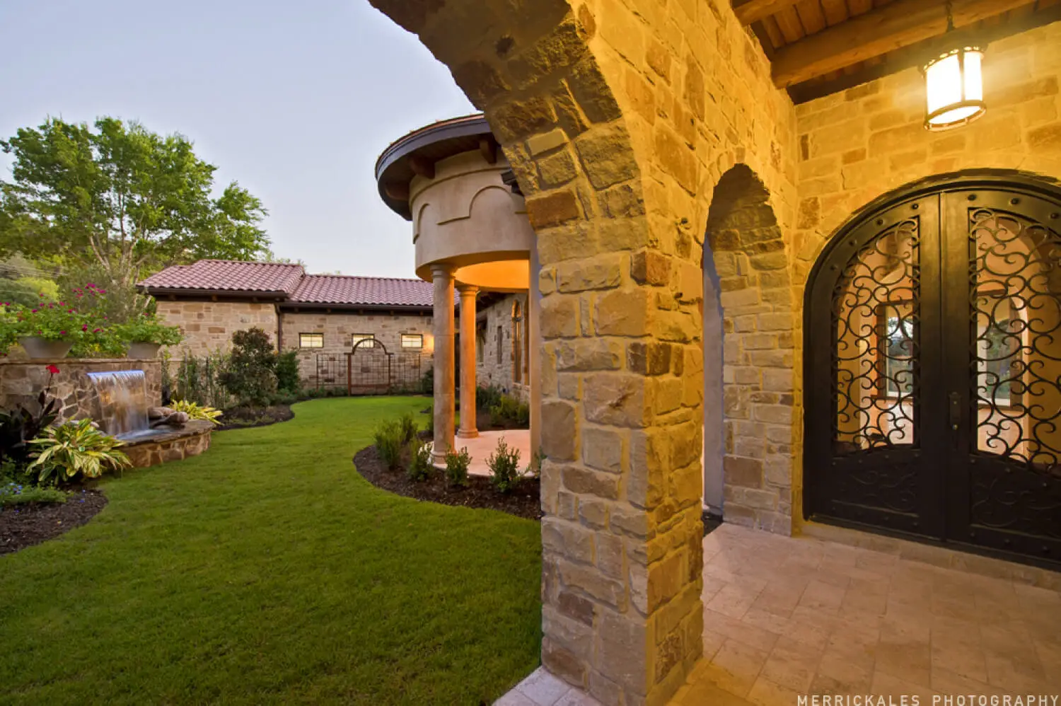 Front elevation at dusk of a stone and stucco home with a tile roof, lit entry arch and a flagstone walk across the lawn.