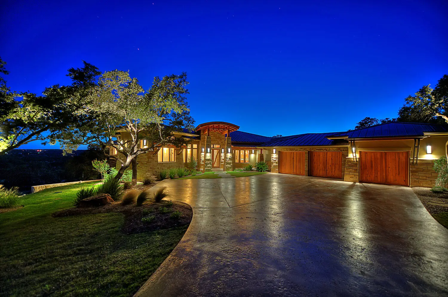 Long low home with a hipped metal roof and stone piers, seen across a lawn under a large live oak.