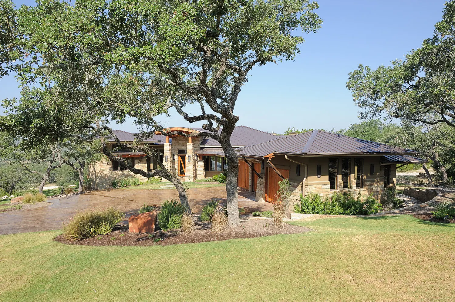Long low house with a hipped metal roof and stone piers, seen across a lawn under a large live oak.